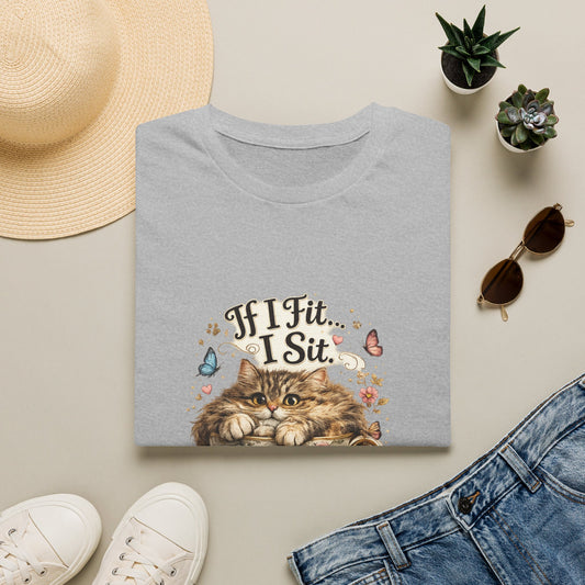 A folded gray graphic T-shirt is displayed in a styled flat lay on a light beige surface. The shirt features a printed design of a fluffy brown cat sitting in a tea cup with the text 'If I fit I sit' surrounded by small colorful floral and butterfly details.