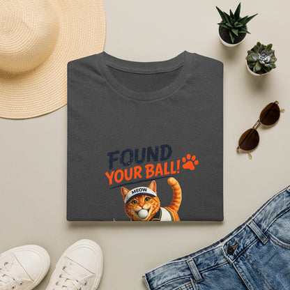 A folded dark charcoal gray t-shirt with a graphic print reading 'FOUND YOUR BALL!' in navy and orange, featuring an orange tabby cat wearing a 'MEOW' cap. The shirt is styled in a flat lay with a straw hat, white sneakers, blue jeans, and small potted succulents on a light beige background.