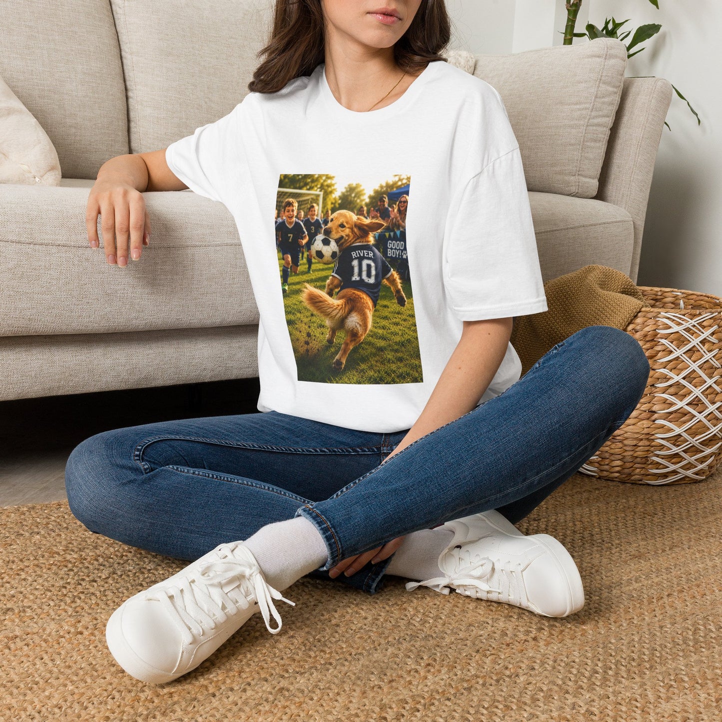 A woman sitting on a woven rug in front of a beige couch, wearing a white graphic t-shirt with a printed photo of a dog playing soccer, blue jeans, and white sneakers.