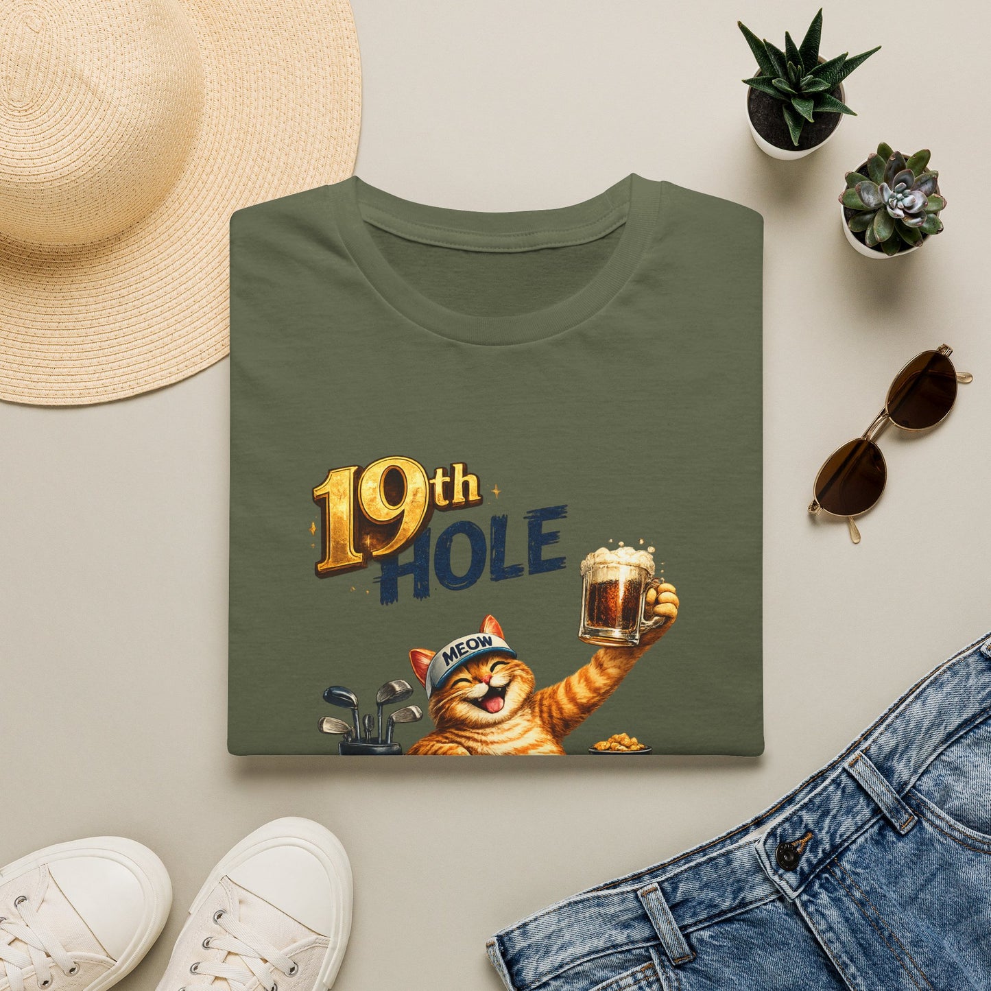 A folded military green graphic T-shirt is arranged in a styled flat lay on a light beige surface. The shirt features a cartoon orange tabby cat wearing a golf cap and holding a mug, with the text '19th HOLE' above it.
