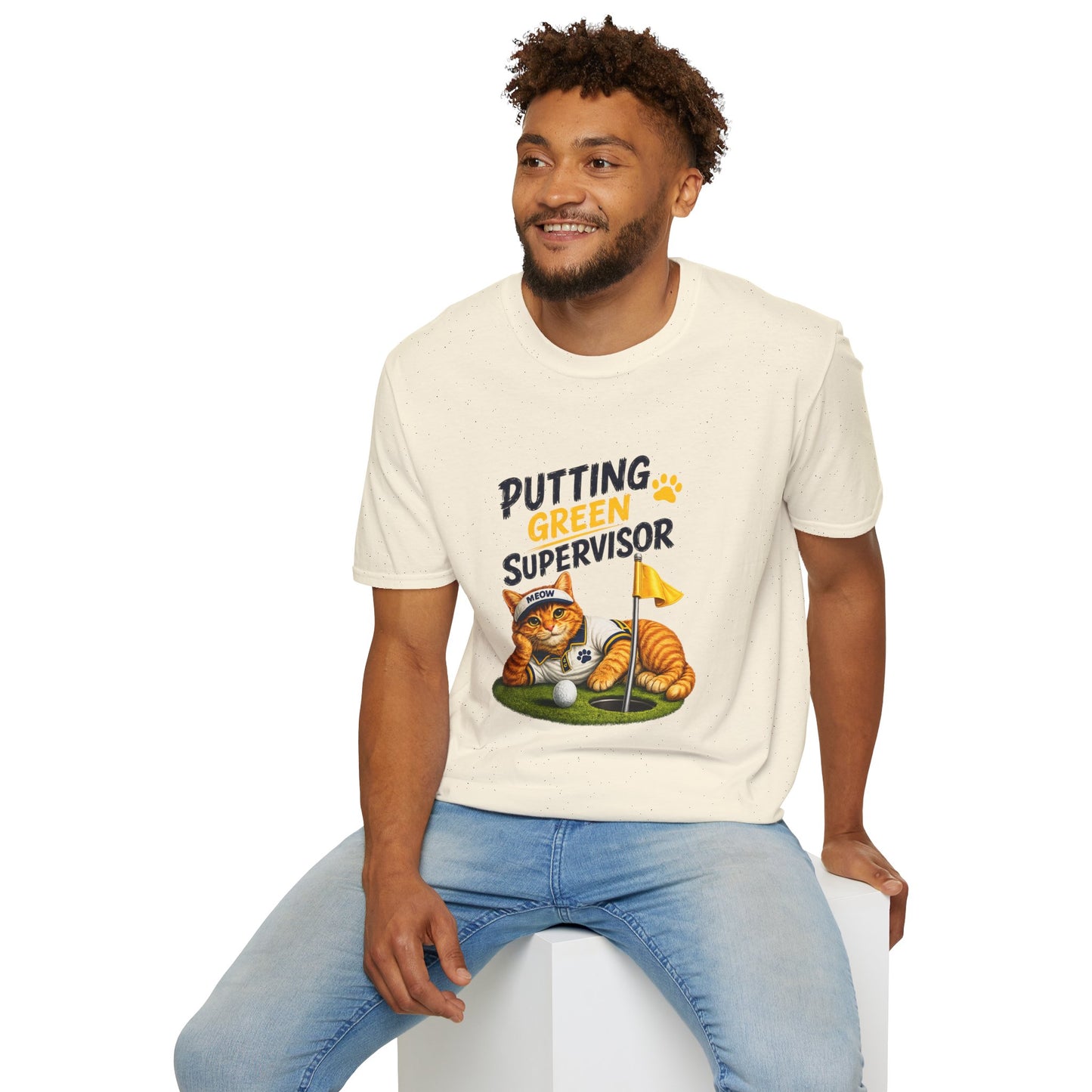 A man wearing a cream-colored graphic t-shirt that reads 'PUTTING GREEN SUPERVISOR' with a cartoon cat on a golf green, paired with light blue jeans, seated on a white block against a white background.