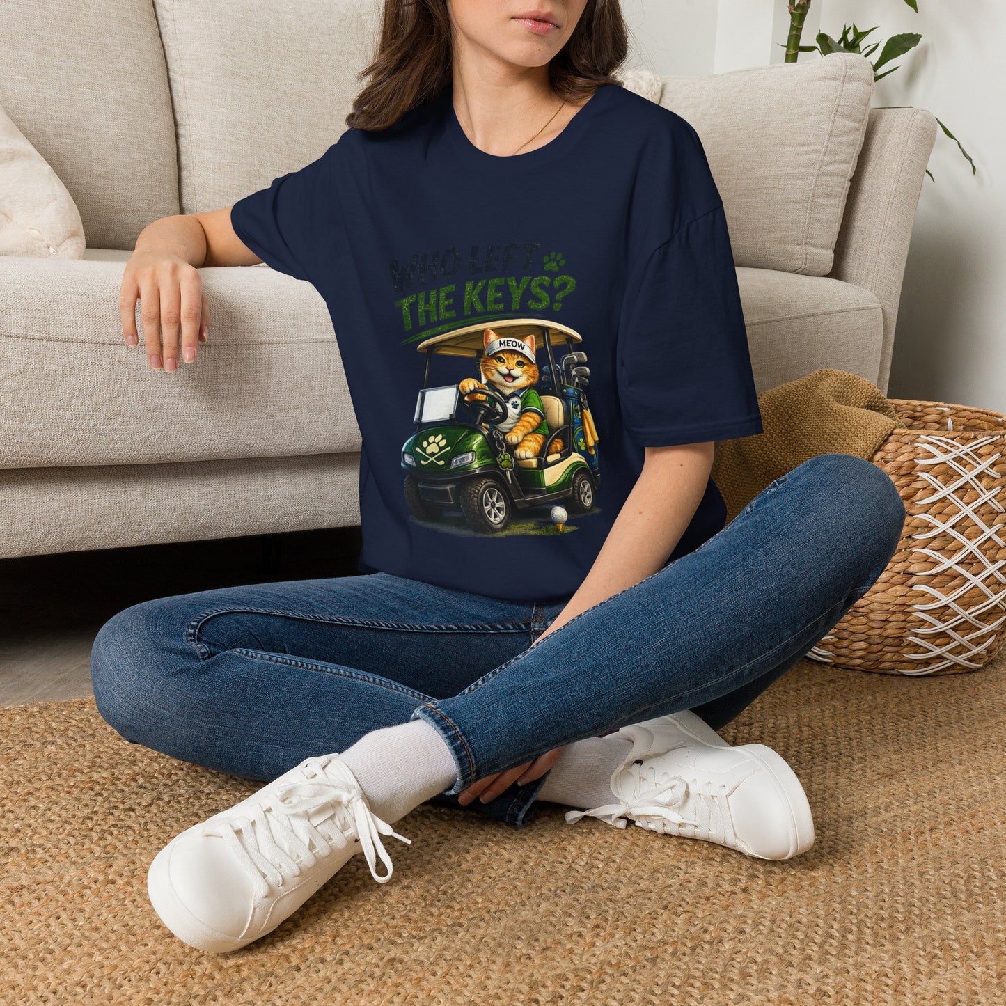 A woman sitting cross-legged on a woven rug in front of a beige sofa, wearing an navy blue graphic T-shirt with a cartoon cat driving a golf cart and the text 'WHO LET THE KEYS?'.
