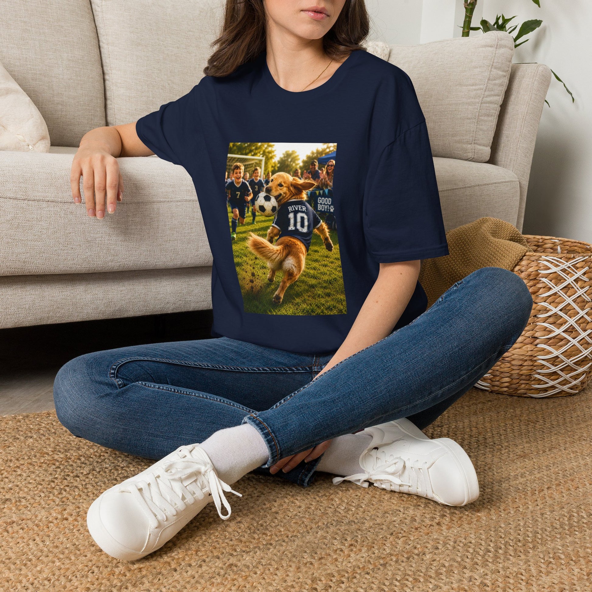 A woman sitting cross-legged on a woven rug in front of a beige sofa, wearing an oversized navy blue graphic T-shirt. The shirt features a rectangular photo print of a golden retriever dog playing fsoccer on a grassy field, wearing a blue jersey with the number 10.
