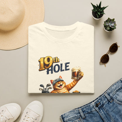 A folded natural color graphic T-shirt is arranged in a styled flat lay on a light beige surface. The shirt features a cartoon orange tabby cat wearing a golf cap and holding a mug, with the text '19th HOLE' above it.
