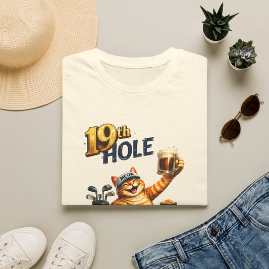 A folded natural color graphic T-shirt is arranged in a styled flat lay on a light beige surface. The shirt features a cartoon orange tabby cat wearing a golf cap and holding a mug, with the text '19th HOLE' above it.