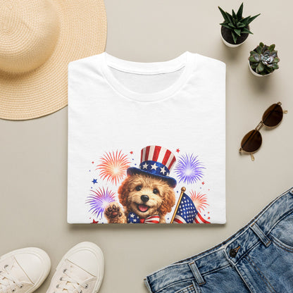 White t-shirt with a dog wearing a patriotic hat and holding an American flag, surrounded by a sun hat, sunglasses, and jeans on a light background.