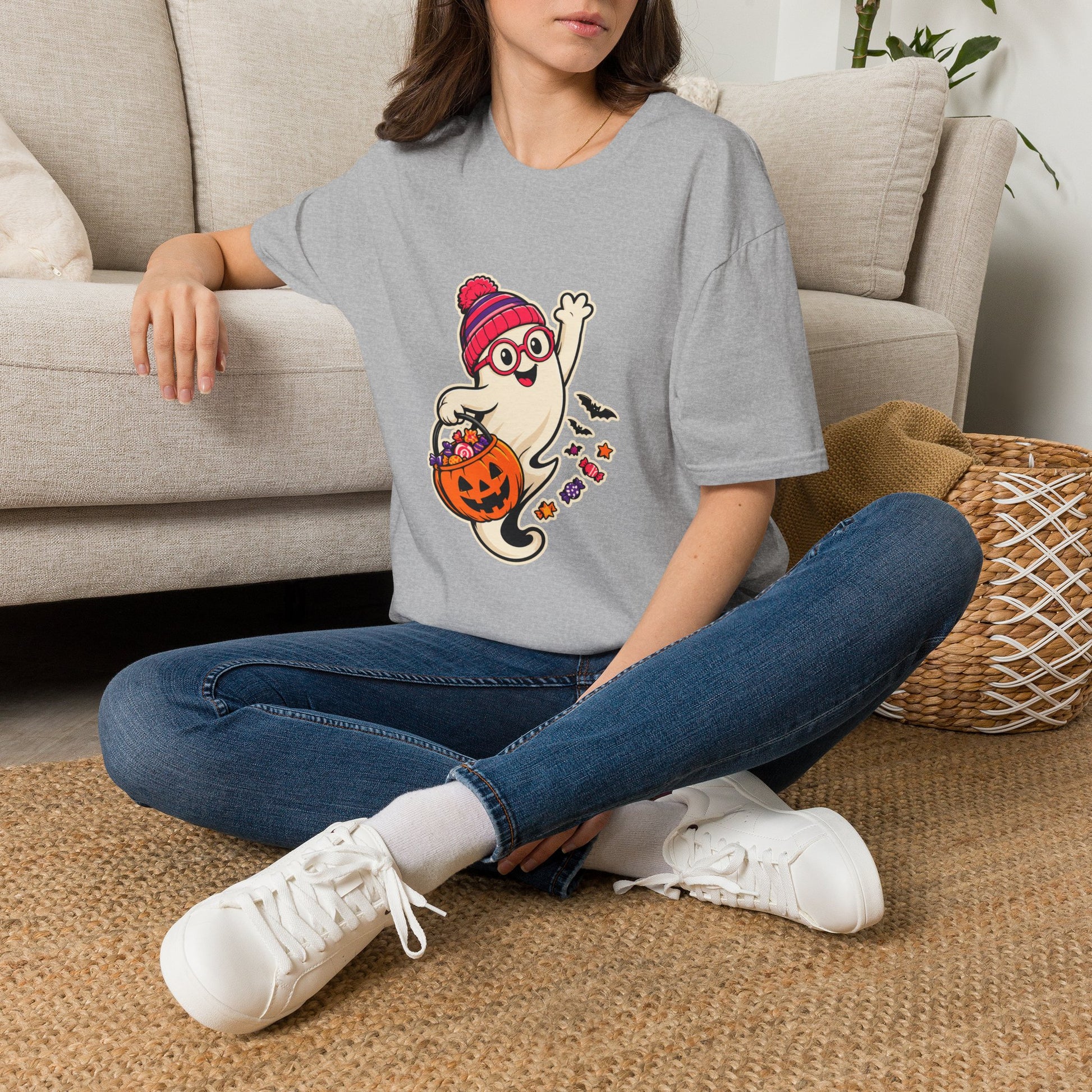 A woman sitting cross-legged on a woven rug in front of a beige sofa, wearing a light gray t-shirt with a cartoon ghost Halloween graphic, blue jeans, and white sneakers.