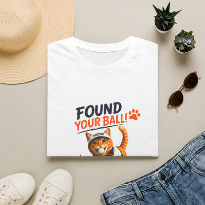 A folded white graphic T-shirt is displayed in a styled flat lay on a light beige surface. The shirt features the text 'FOUND YOUR BALL!' in black and orange lettering with an orange paw print, above an illustration of an orange tabby cat wearing a 'MEOW' bandana.