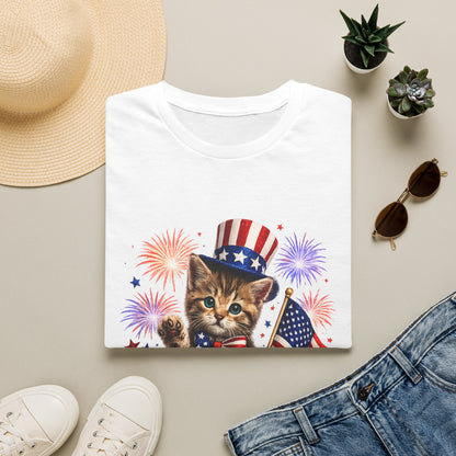 White t-shirt with a kitten wearing an American flag hat design, surrounded by a straw hat, sunglasses, sneakers, and jeans on a beige surface.