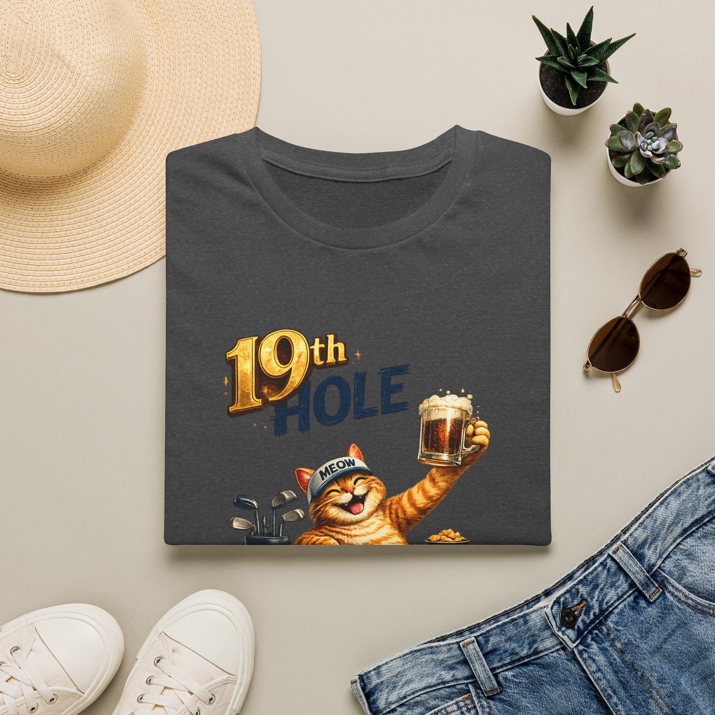 A folded dark heather graphic T-shirt is arranged in a styled flat lay on a light beige surface. The shirt features a cartoon orange tabby cat wearing a golf cap and holding a mug, with the text '19th HOLE' above it.
