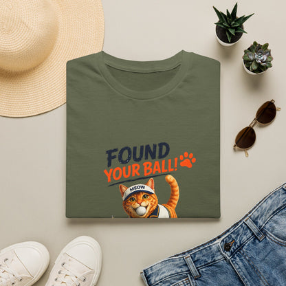 A foldedmilitary green T-shirt with a humorous graphic print reading 'FOUND YOUR BALL!' in dark blue and orange text, featuring an orange tabby cat wearing a headband that says 'MEOW'. The shirt is styled in a flat-lay arrangement with a straw hat, white sneakers, blue jeans, sunglasses, and small potted succulents on a light beige background.