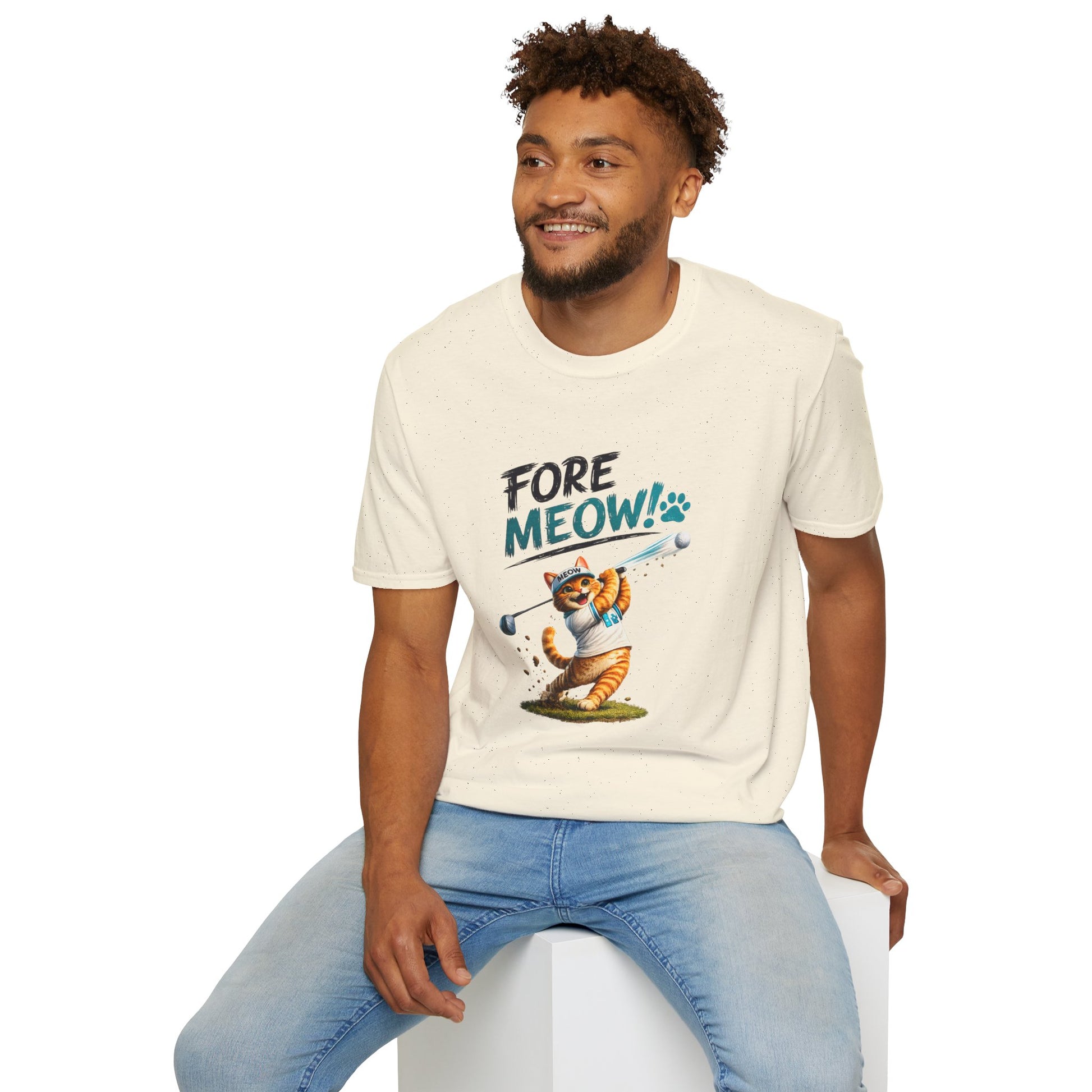 A man wearing a cream-colored graphic t-shirt with a cartoon cat illustration and the text 'FORE MEOW!'. He is sitting on a white block against a plain white background.