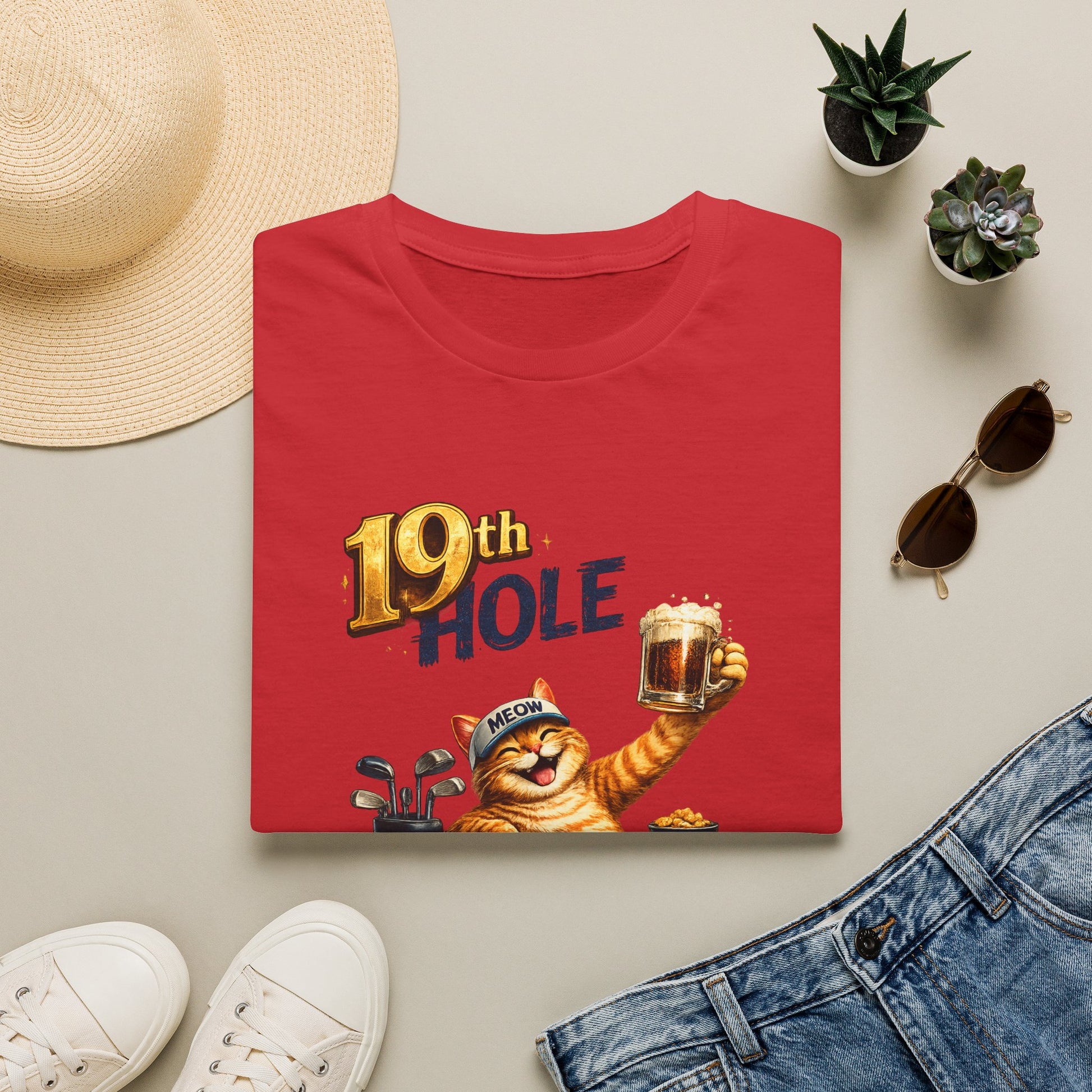 A folded red graphic T-shirt is arranged in a styled flat lay on a light beige surface. The shirt features a cartoon orange tabby cat wearing a golf cap and holding a mug, with the text '19th HOLE' above it.