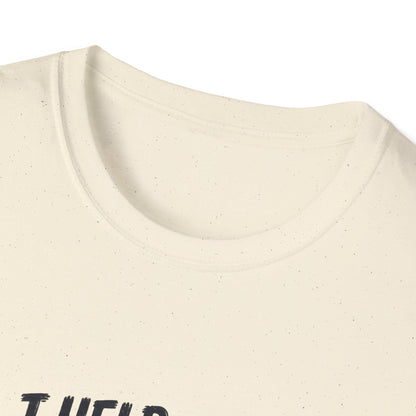 A folded natural color graphic T-shirt collar is displayed in a styled flat lay on a light beige surface. The shirt features a cartoon cat wearing a baseball cap and holding golf clubs, with the text 'I HELP WITH YOUR SHORT GAME BY STEALING GOLF BALLS.