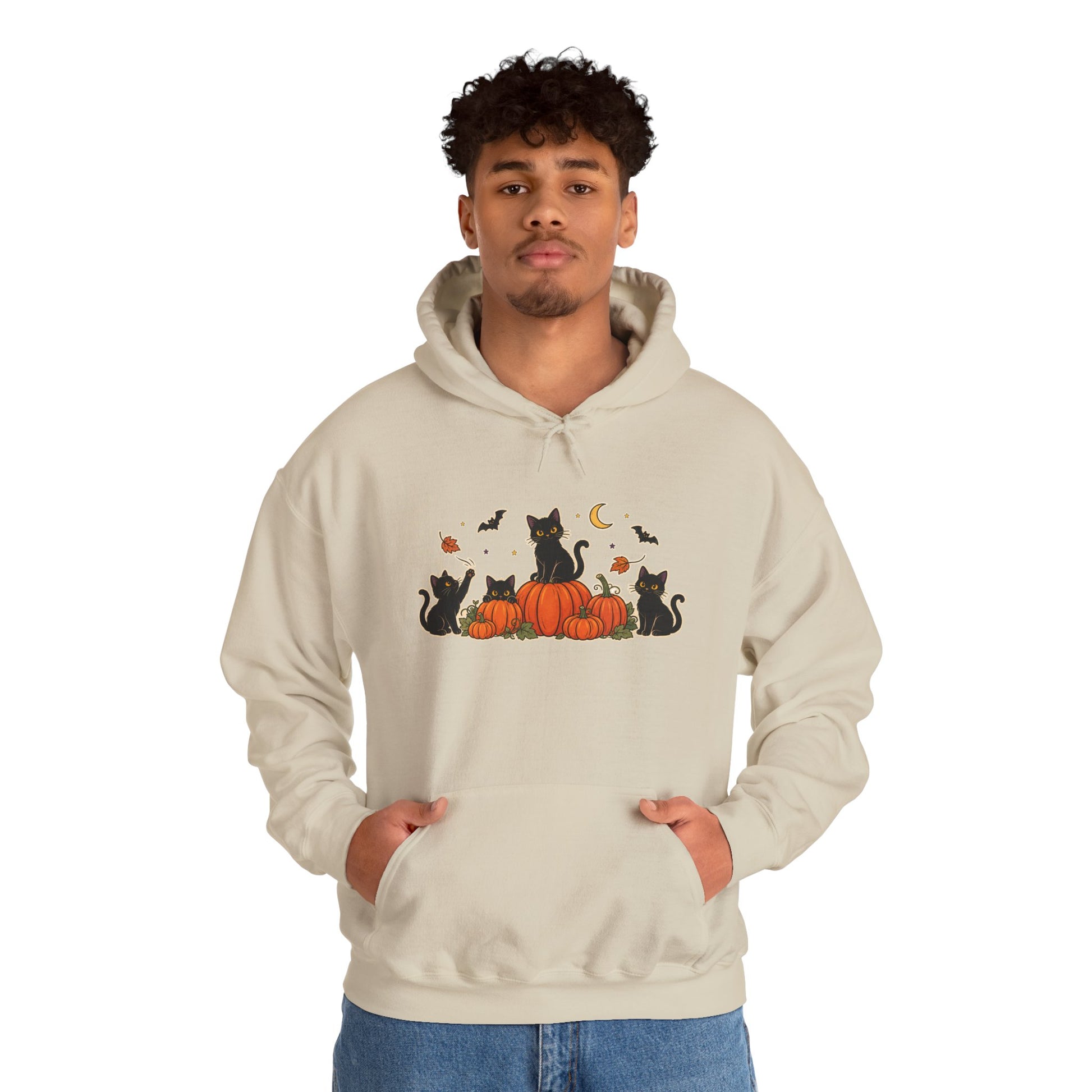 A man wearing a light beige hoodie with a graphic print of black cats sitting on orange pumpkins with bats, a crescent moon, and acorns. He is standing against a plain white background.