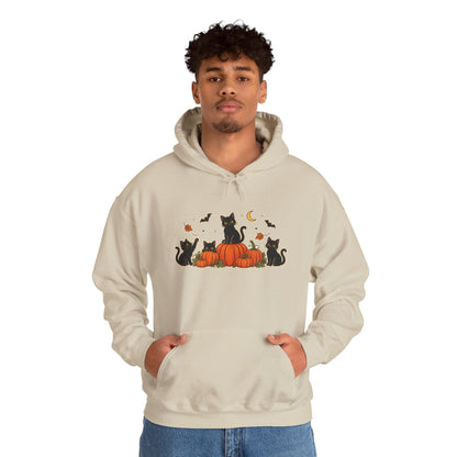 A man wearing a light beige hoodie with a graphic print of black cats sitting on orange pumpkins with bats, a crescent moon, and acorns. He is standing against a plain white background.