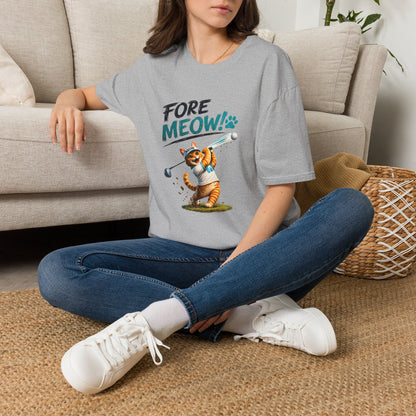 A woman sitting cross-legged on a woven rug in front of a beige sofa, wearing a light gray graphic T-shirt with the text 'FORE MEOW!' and a cartoon tiger holding a golf club. She is also wearing blue jeans and white sneakers.