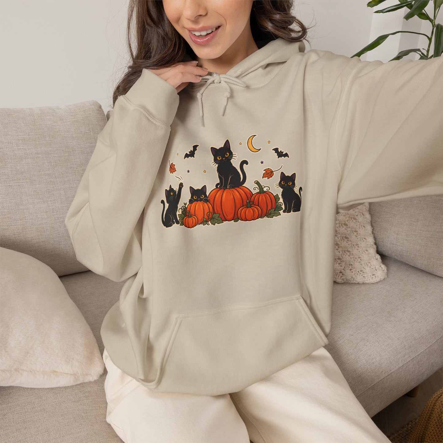 A woman wearing a beige hoodie with a graphic of black cats around orange pumpkins and small bats, sitting on a light gray couch.