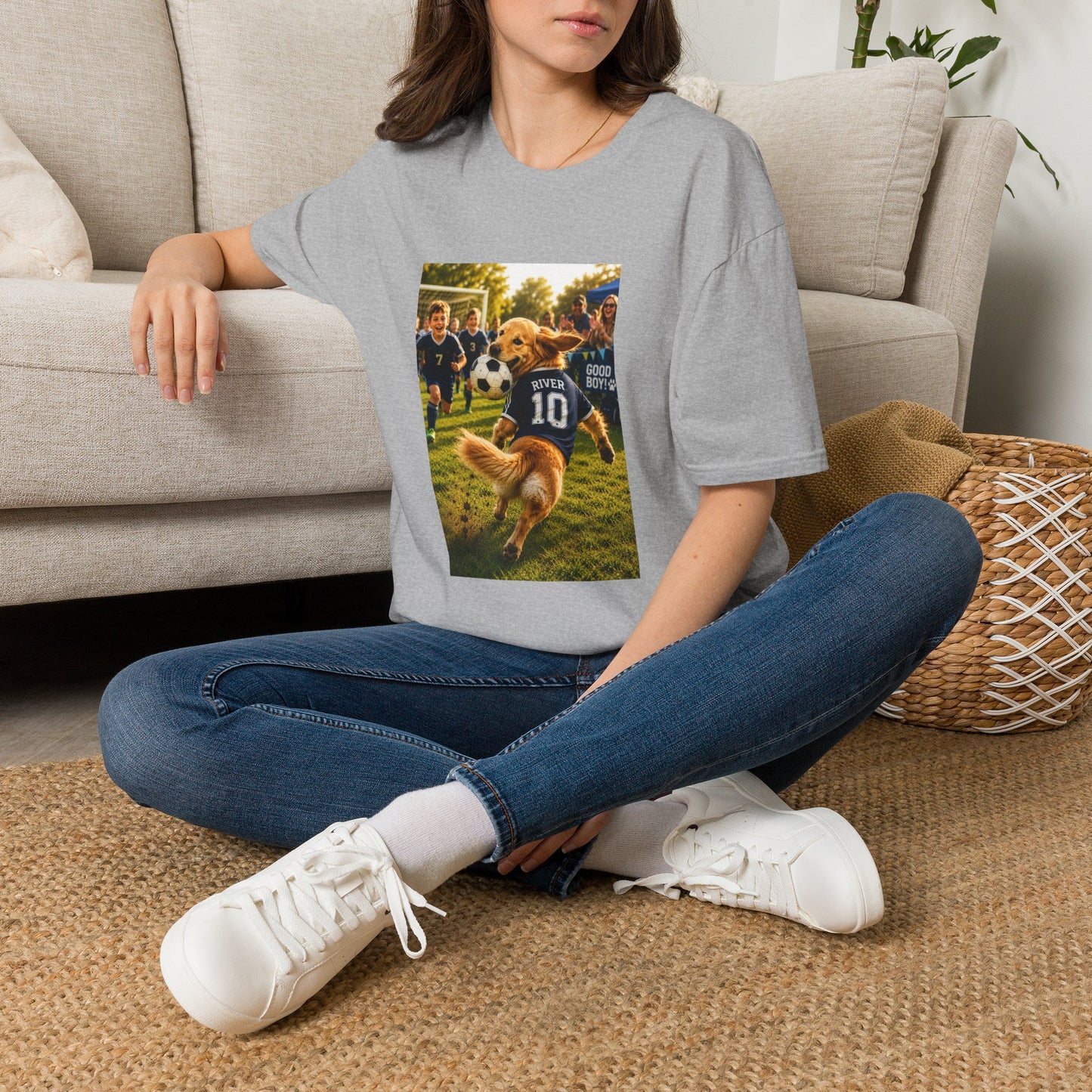 A woman sitting cross-legged on a woven rug in front of a beige sofa, wearing a light gray t-shirt with a custom graphic print of a dog playing soccer. The shirt features a photo of a golden retriever dog wearing a blue 'PUPPET 10' jersey on a grassy field.