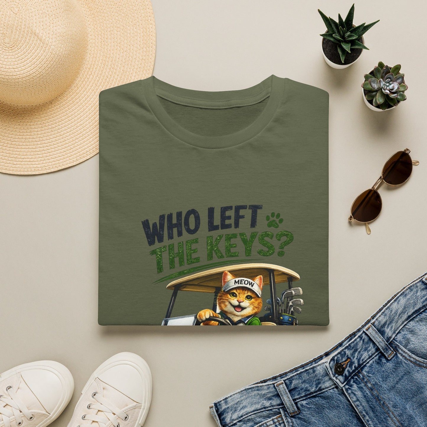 An olive green t-shirt with a graphic of a cat driving a golf cart and the text 'WHO LEFT THE KEYS?' is displayed in a flat-lay arrangement with a straw hat, white sneakers, blue jeans, and small potted succulents on a light beige background.