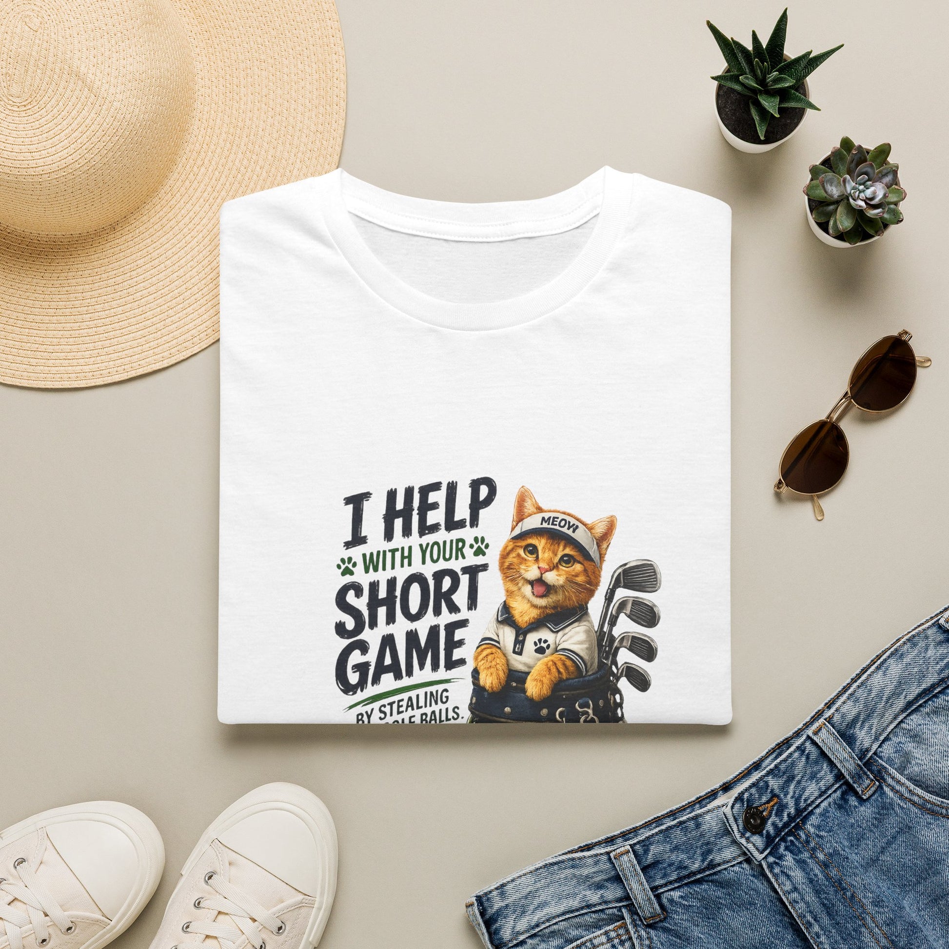 A folded white graphic T-shirt is displayed in a styled flat lay on a light beige surface. The shirt features a cartoon cat wearing a baseball cap and holding golf clubs, with the text 'I HELP WITH YOUR SHORT GAME BY STEALING GOLF BALLS.