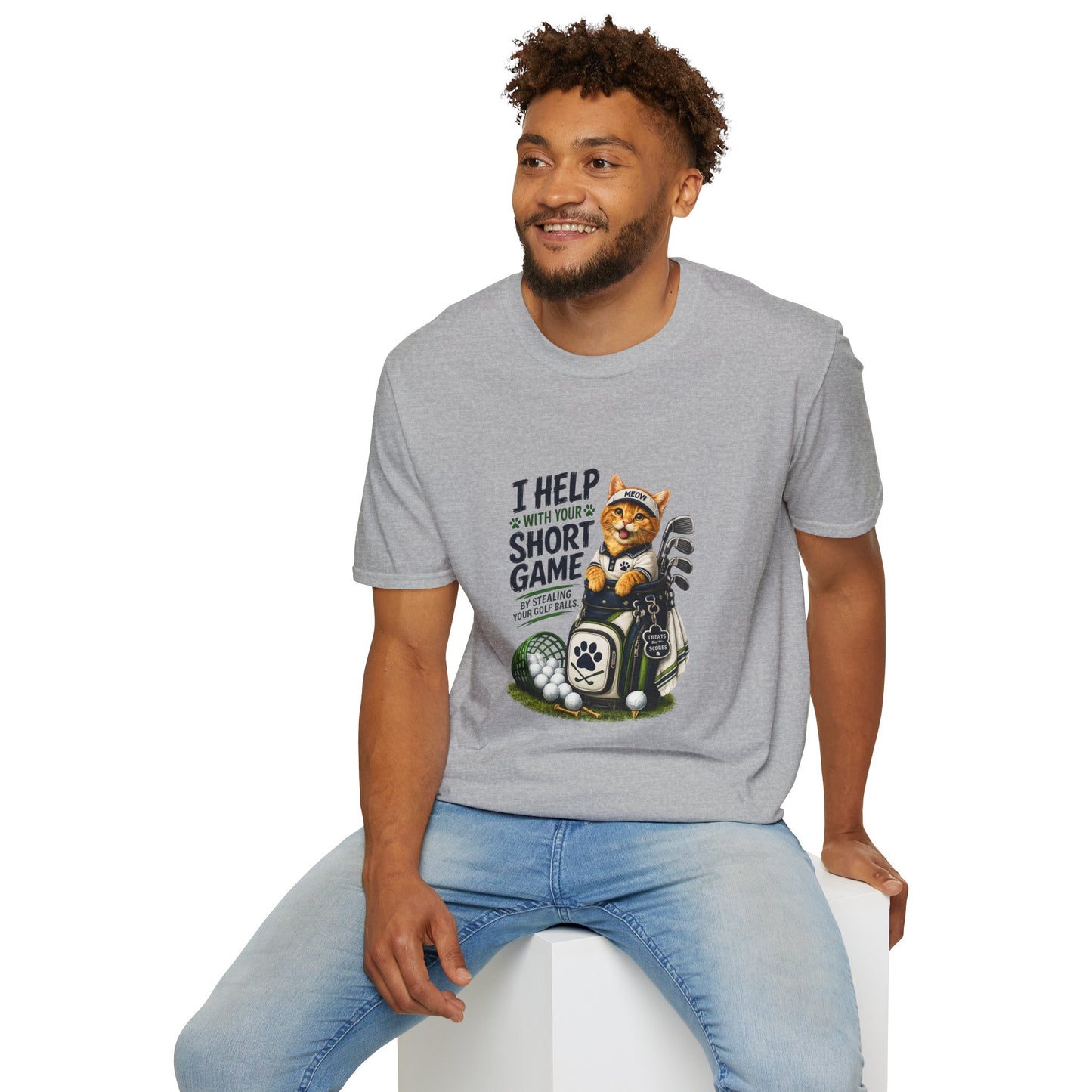 A sport grey graphic T-shirt is displayed in a styled flat lay on a light beige surface. The shirt features a cartoon cat wearing a baseball cap and holding golf clubs, with the text 'I HELP WITH YOUR SHORT GAME BY STEALING GOLF BALLS.