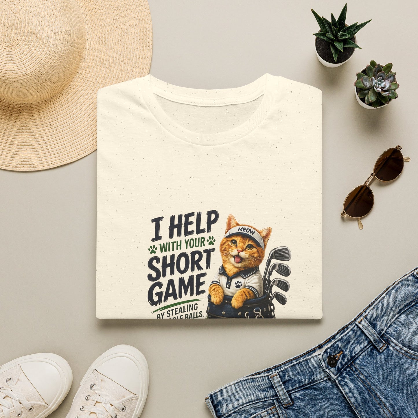 A folded natural graphic T-shirt is displayed in a styled flat lay on a light beige surface. The shirt features a cartoon cat wearing a baseball cap and holding golf clubs, with the text 'I HELP WITH YOUR SHORT GAME BY STEALING GOLF BALLS.