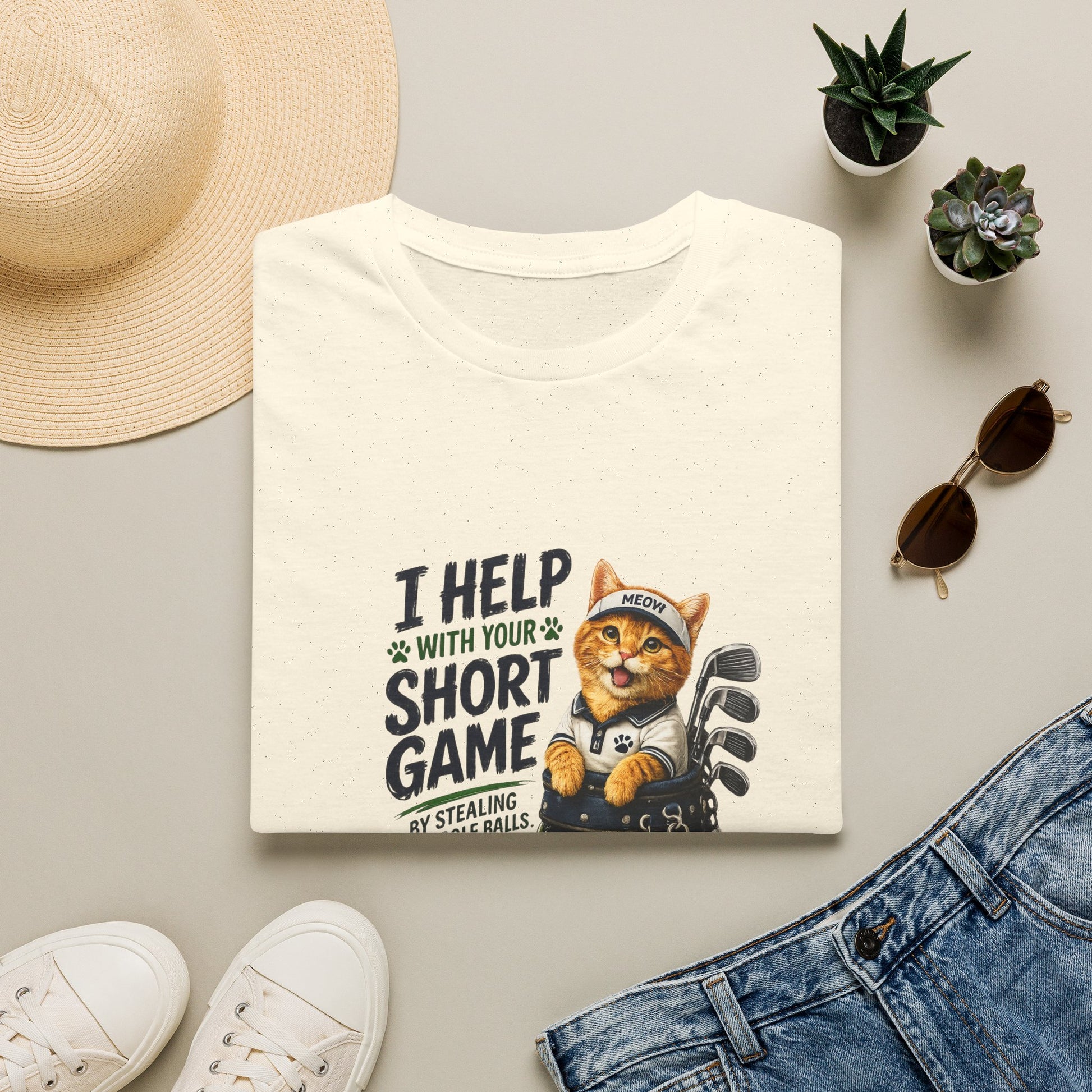 A folded natural graphic T-shirt is displayed in a styled flat lay on a light beige surface. The shirt features a cartoon cat wearing a baseball cap and holding golf clubs, with the text 'I HELP WITH YOUR SHORT GAME BY STEALING GOLF BALLS.