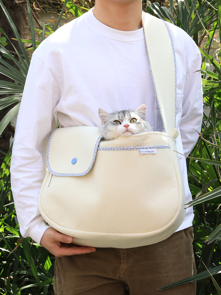 Person holding a beige pet carrier with a cat peeking out, surrounded by green foliage.