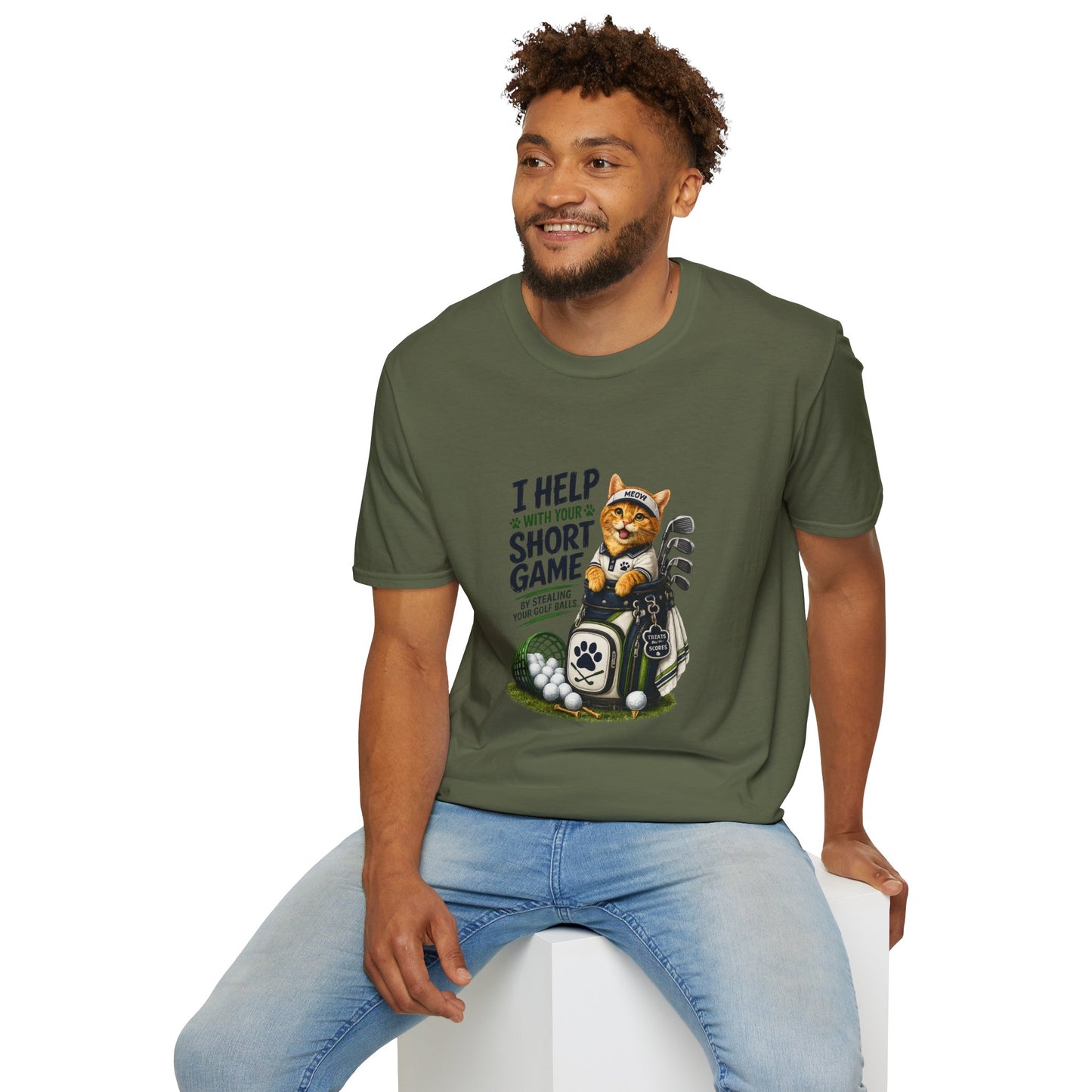 A military green graphic T-shirt is displayed in a styled flat lay on a light beige surface. The shirt features a cartoon cat wearing a baseball cap and holding golf clubs, with the text 'I HELP WITH YOUR SHORT GAME BY STEALING GOLF BALLS.