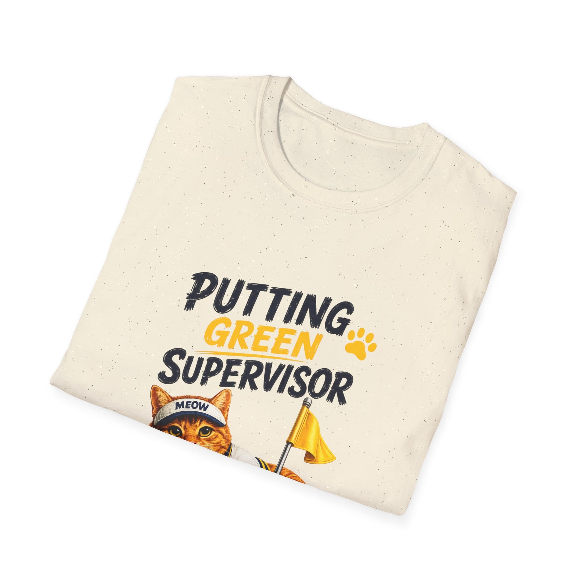 A folded cream-colored t-shirt with a graphic print that reads 'PUTTING GREEN SUPERVISOR' in black and yellow text, featuring an illustration of an orange tabby cat wearing a 'MEOW' cap and holding a yellow flag.