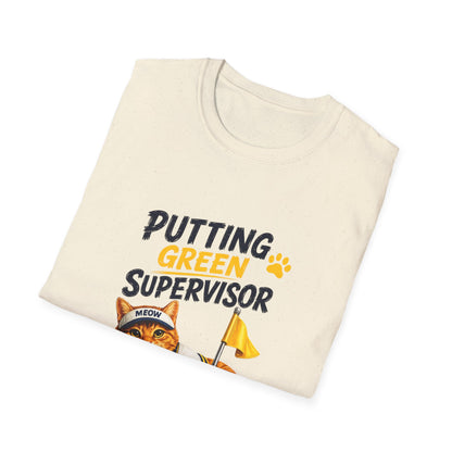A folded cream-colored t-shirt with a graphic print that reads 'PUTTING GREEN SUPERVISOR' in black and yellow text, featuring an illustration of an orange tabby cat wearing a 'MEOW' cap and holding a yellow flag.