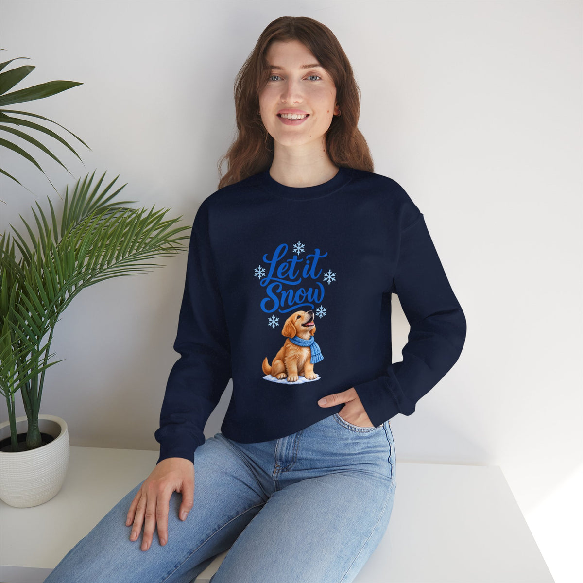 A woman wearing a navy blue crewneck sweatshirt with a graphic of a golden retriever dog in a winter outfit and the text 'Let it Snow' in blue.  