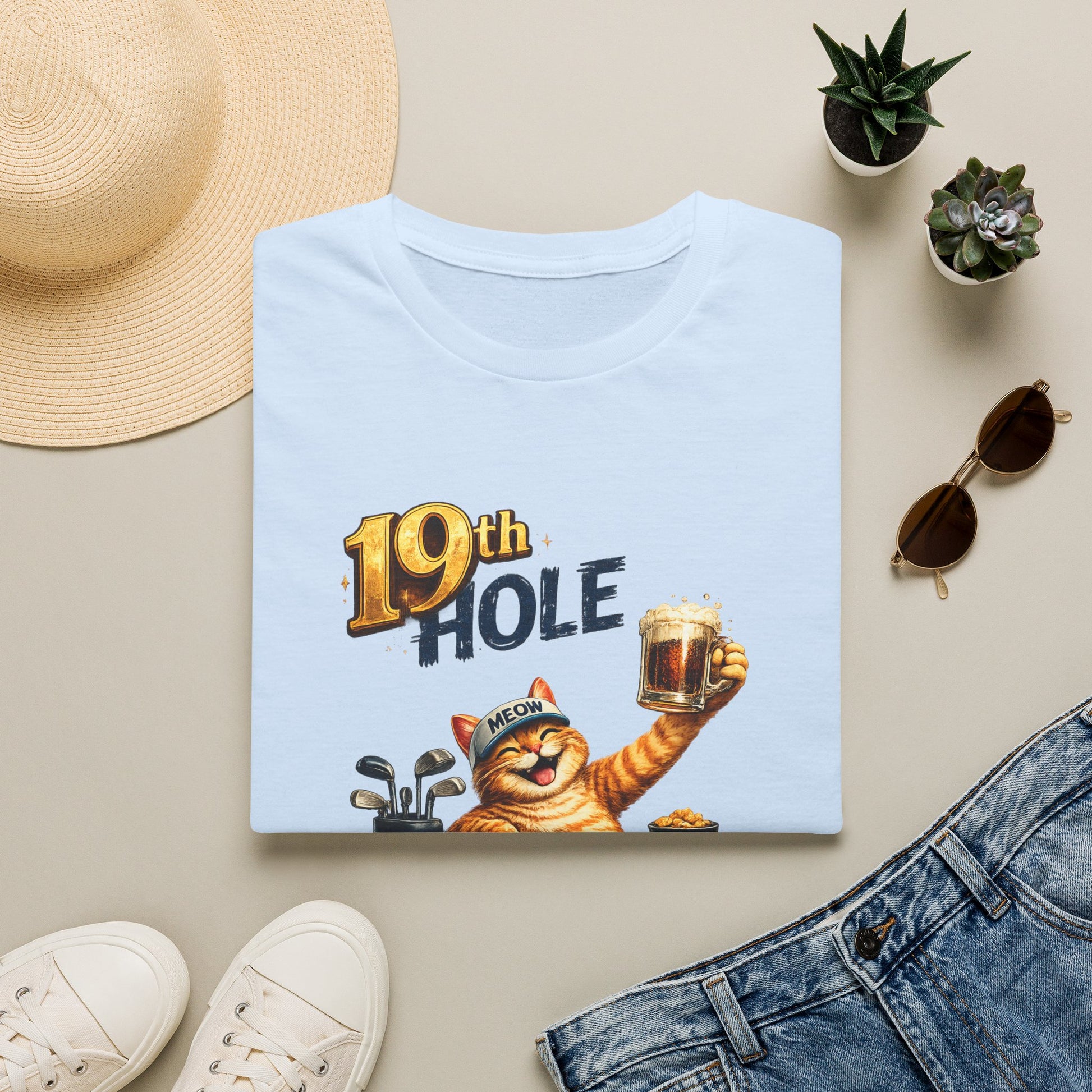 A folded light blue graphic T-shirt is arranged in a styled flat lay on a light beige surface. The shirt features a cartoon orange tabby cat wearing a golf cap and holding a mug, with the text '19th HOLE' above it.