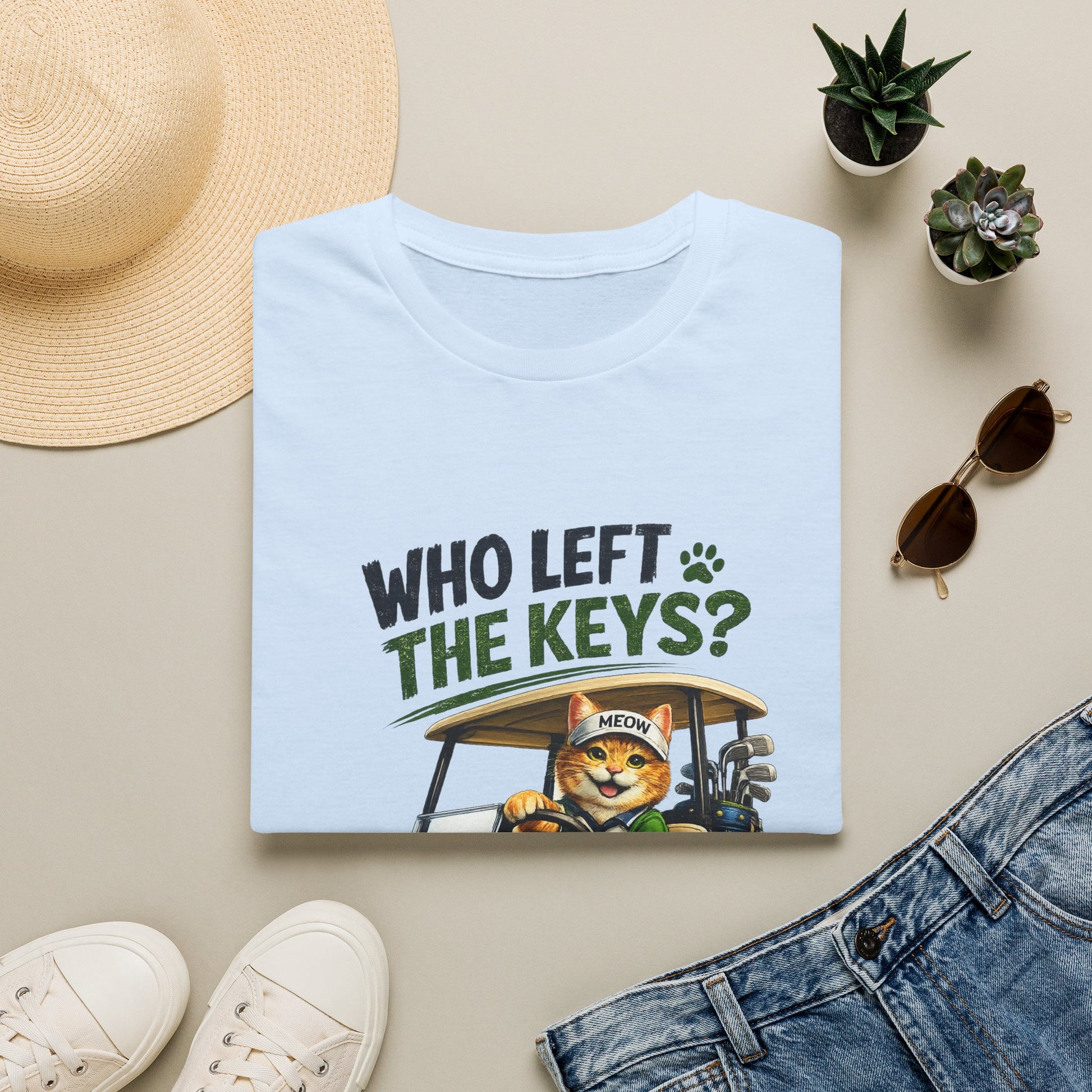 A folded light blue T-shirt with a graphic print reading 'WHO LEFT THE KEYS?' and showing a cartoon cat driving a golf cart, styled in a flat lay with a straw hat, sunglasses, small potted succulents, white sneakers, and blue jeans on a beige background.
