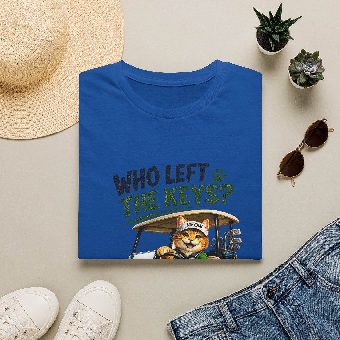 A folded blue graphic T-shirt is displayed in a styled flat lay on a light beige surface. The shirt features a cartoon cat driving a golf cart with the text 'WHO LEFT THE KEYS?' and a paw print.