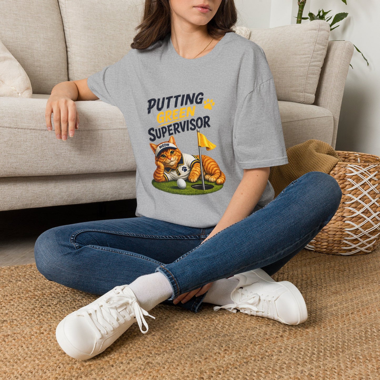 A woman sitting cross-legged on a woven rug in front of a beige sofa, wearing a light gray graphic T-shirt that reads 'PUTTING GREEN SUPERVISOR' with an illustration of an orange tabby cat and a golf ball.