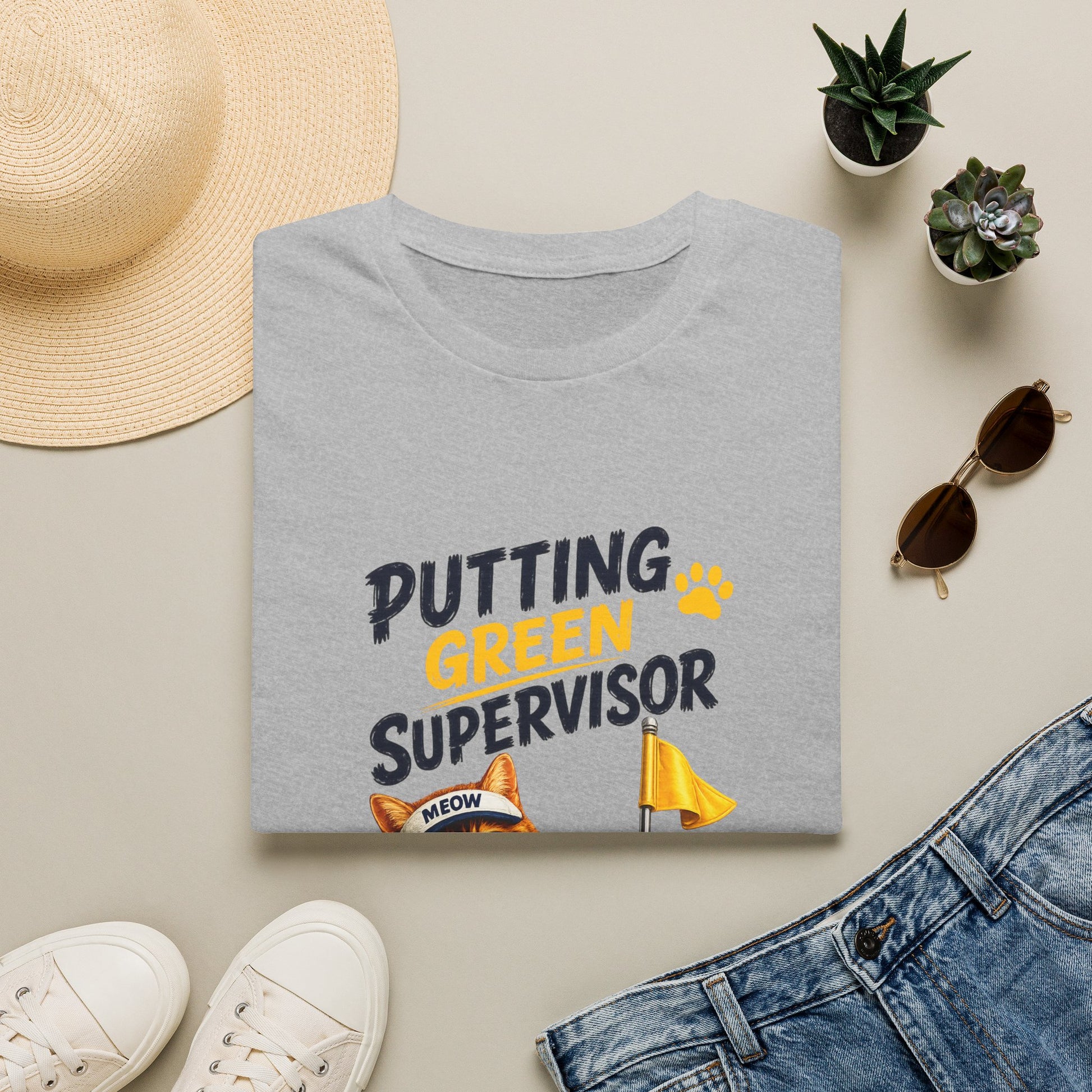 A folded heather gray graphic T-shirt is styled in a flat lay with a straw hat, white sneakers, blue jeans, sunglasses, and small potted succulents. The shirt reads 'PUTTING GREEN SUPERVISOR' in dark gray and yellow text with a small orange cat graphic and yellow flag illustration.