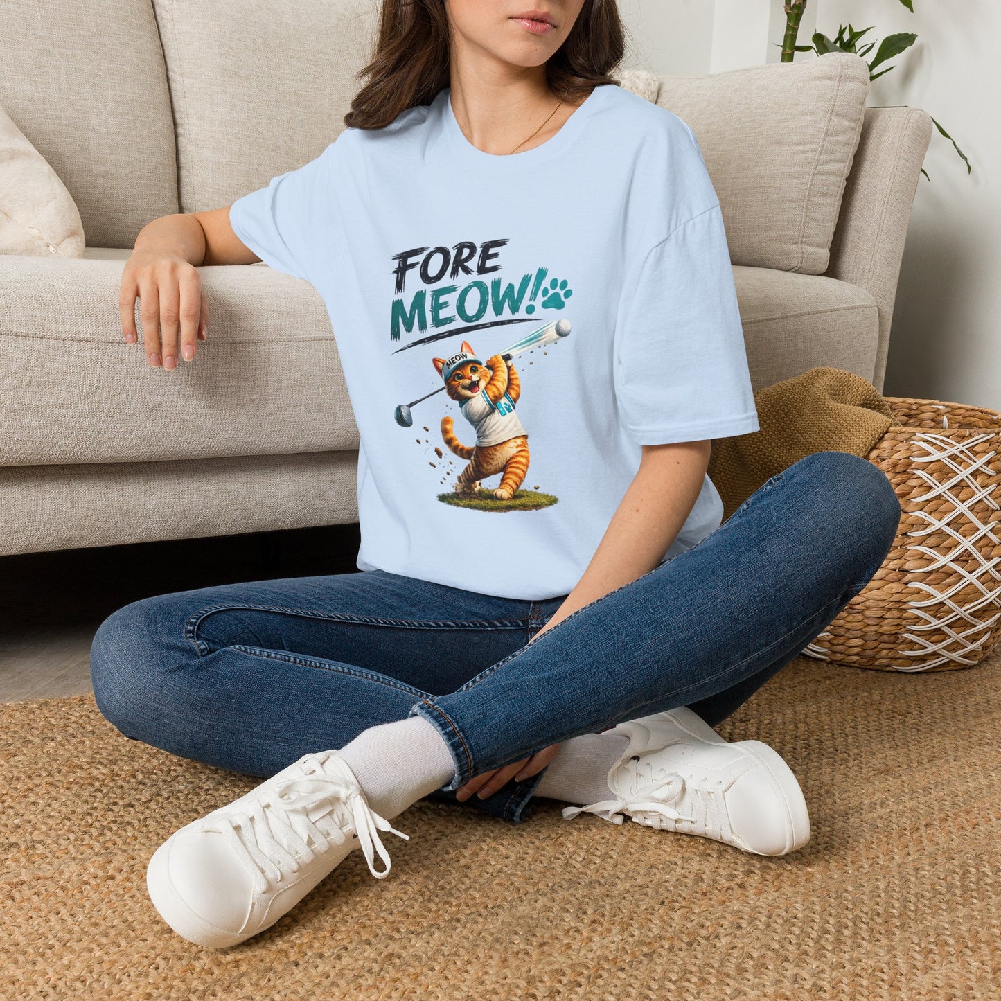 A woman sitting on a woven rug in front of a beige sofa, wearing a light blue graphic T-shirt with the text 'FORE MEOW!' and a cartoon cat holding a golf club, paired with blue jeans and white sneakers.