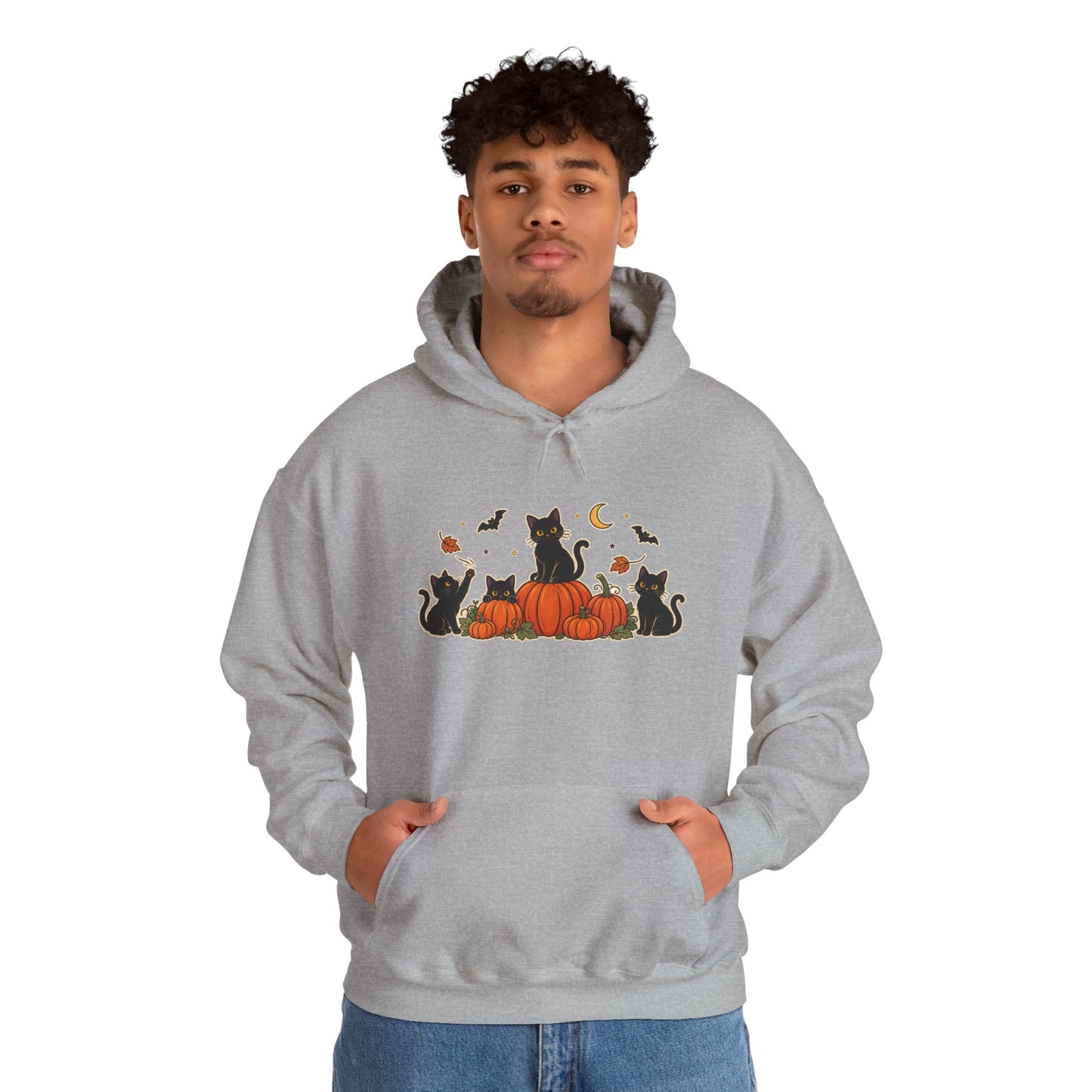 A man wearing a light gray pullover hoodie with a graphic of black cats sitting on orange pumpkins under a crescent moon and bats. The man is also wearing blue jeans, set against a plain white background.