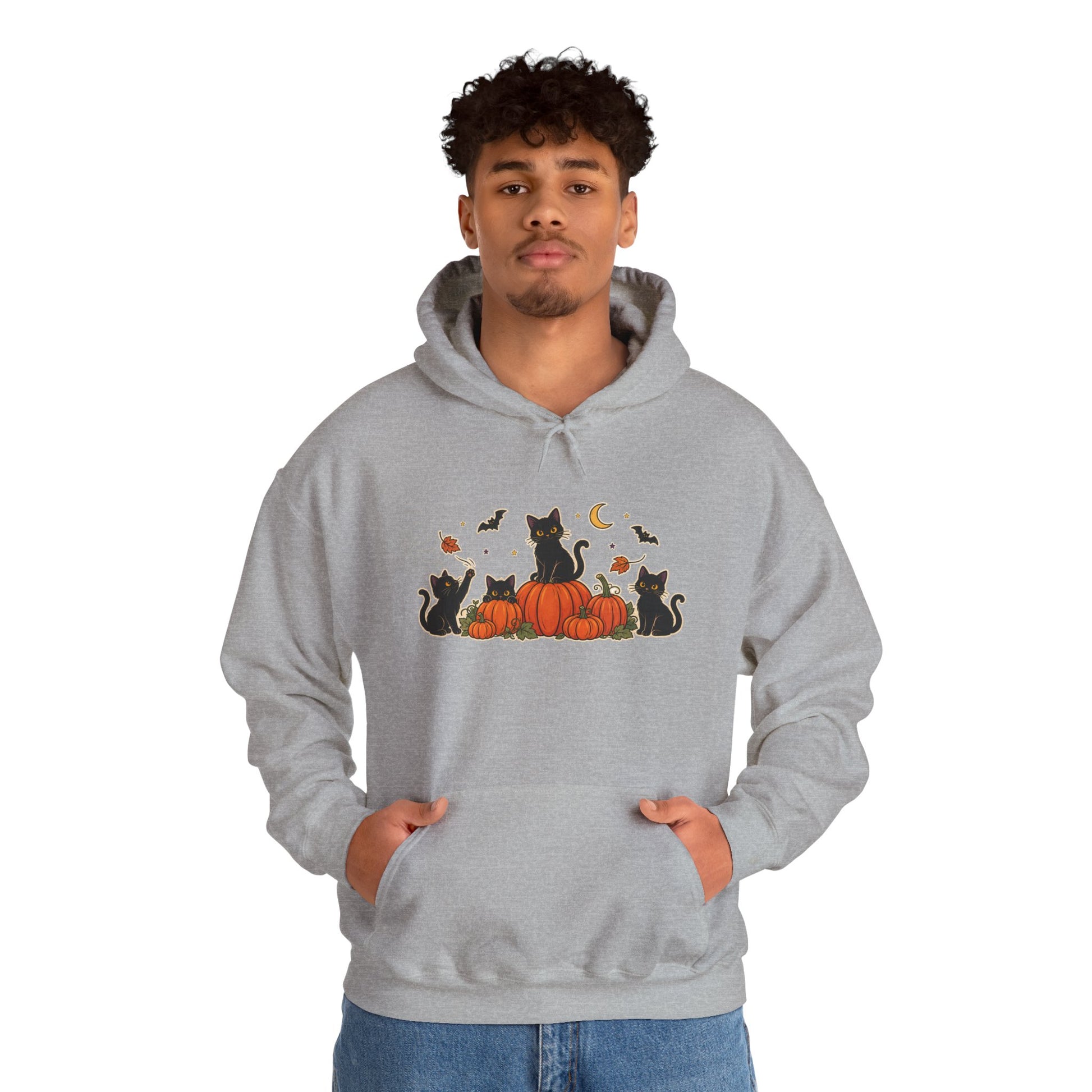 A man wearing a light gray pullover hoodie with a graphic of black cats sitting on orange pumpkins under a crescent moon and bats. The man is also wearing blue jeans, set against a plain white background.