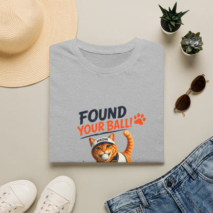 A folded light gray T-shirt with a humorous graphic print reading 'FOUND YOUR BALL!' in dark blue and orange text, featuring an orange tabby cat wearing a headband that says 'MEOW'. The shirt is styled in a flat-lay arrangement with a straw hat, white sneakers, blue jeans, sunglasses, and small potted succulents on a light beige background.