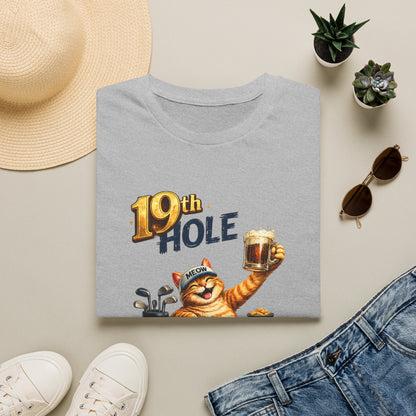A folded sport grey graphic T-shirt is arranged in a styled flat lay on a light beige surface. The shirt features a cartoon orange tabby cat wearing a golf cap and holding a mug, with the text '19th HOLE' above it.