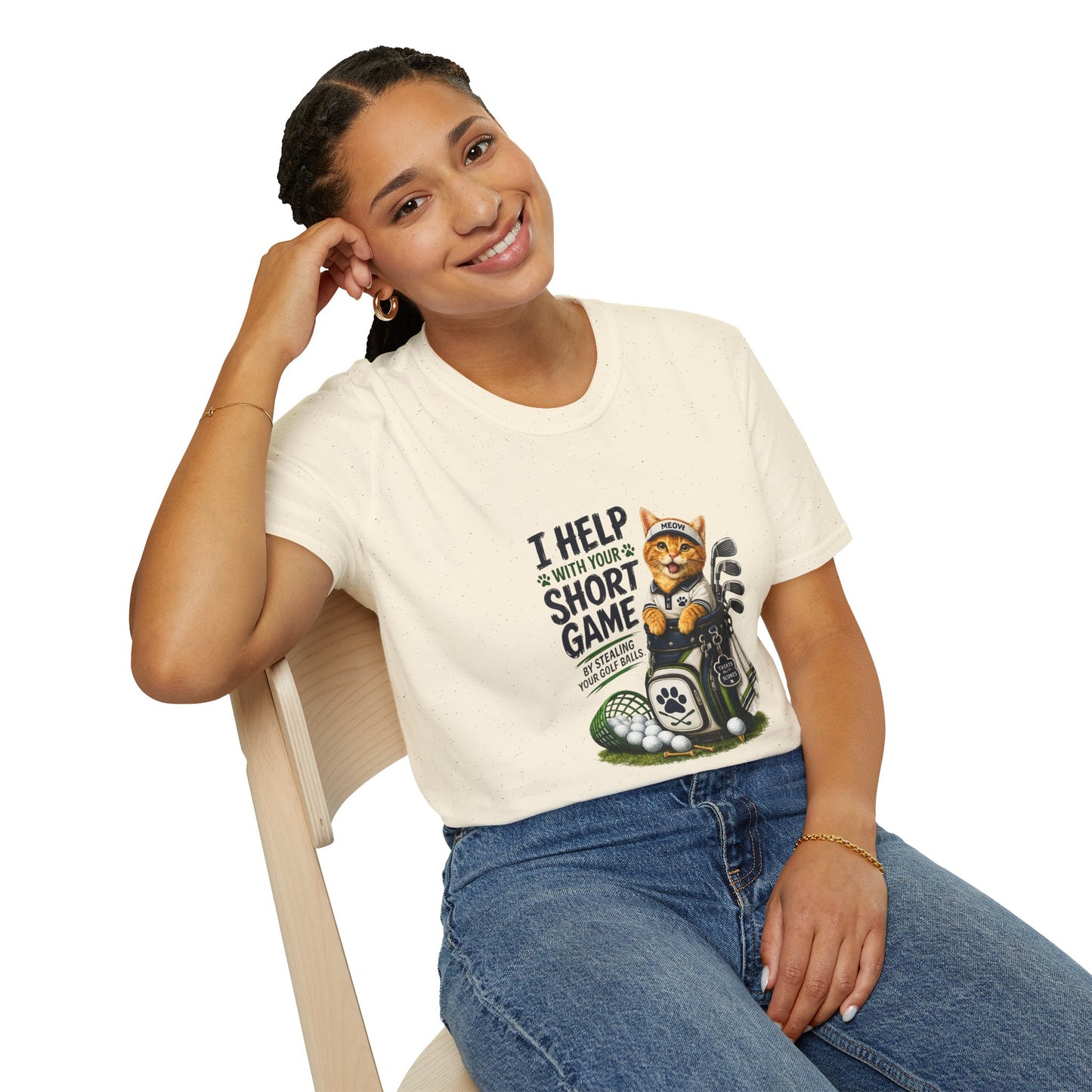 A natural color graphic T-shirt is displayed in a styled flat lay on a light beige surface. The shirt features a cartoon cat wearing a baseball cap and holding golf clubs, with the text 'I HELP WITH YOUR SHORT GAME BY STEALING GOLF BALLS.