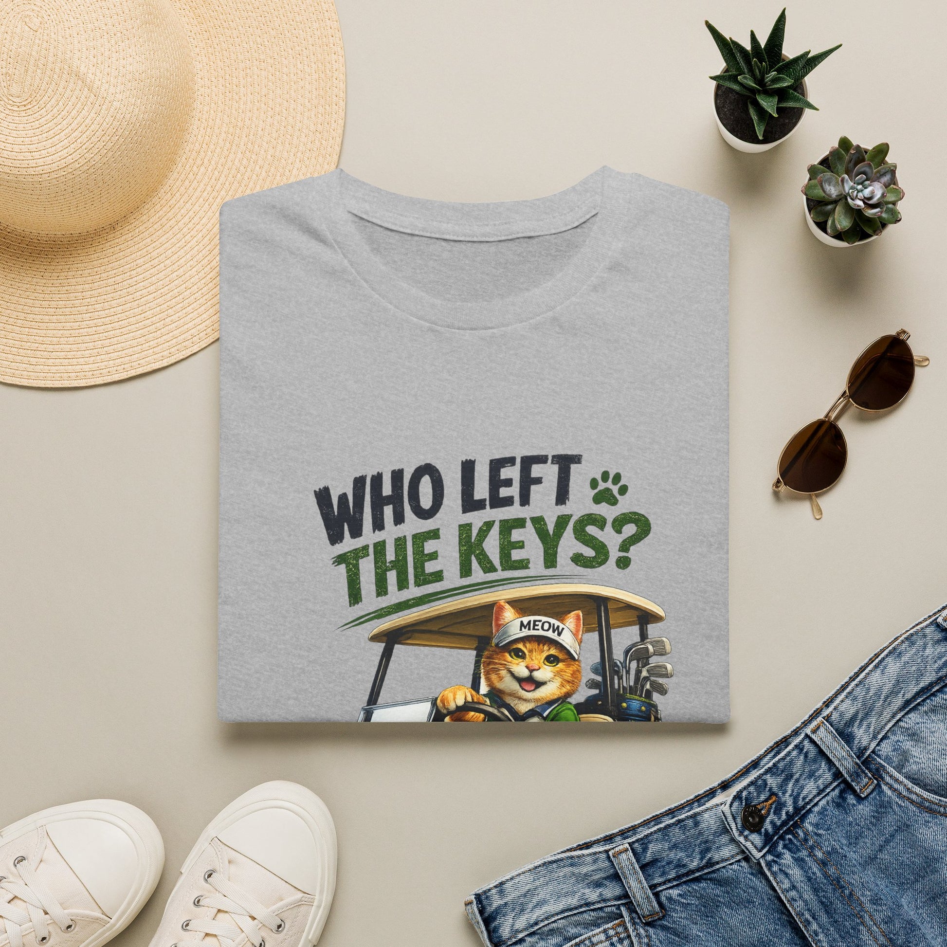 A folded heather gray T-shirt with a graphic print reading 'WHO LEFT THE KEYS?' and showing an orange cat driving a golf cart, styled in a flat lay with a straw hat, white sneakers, sunglasses, small potted succulents, and blue jeans on a light beige background.