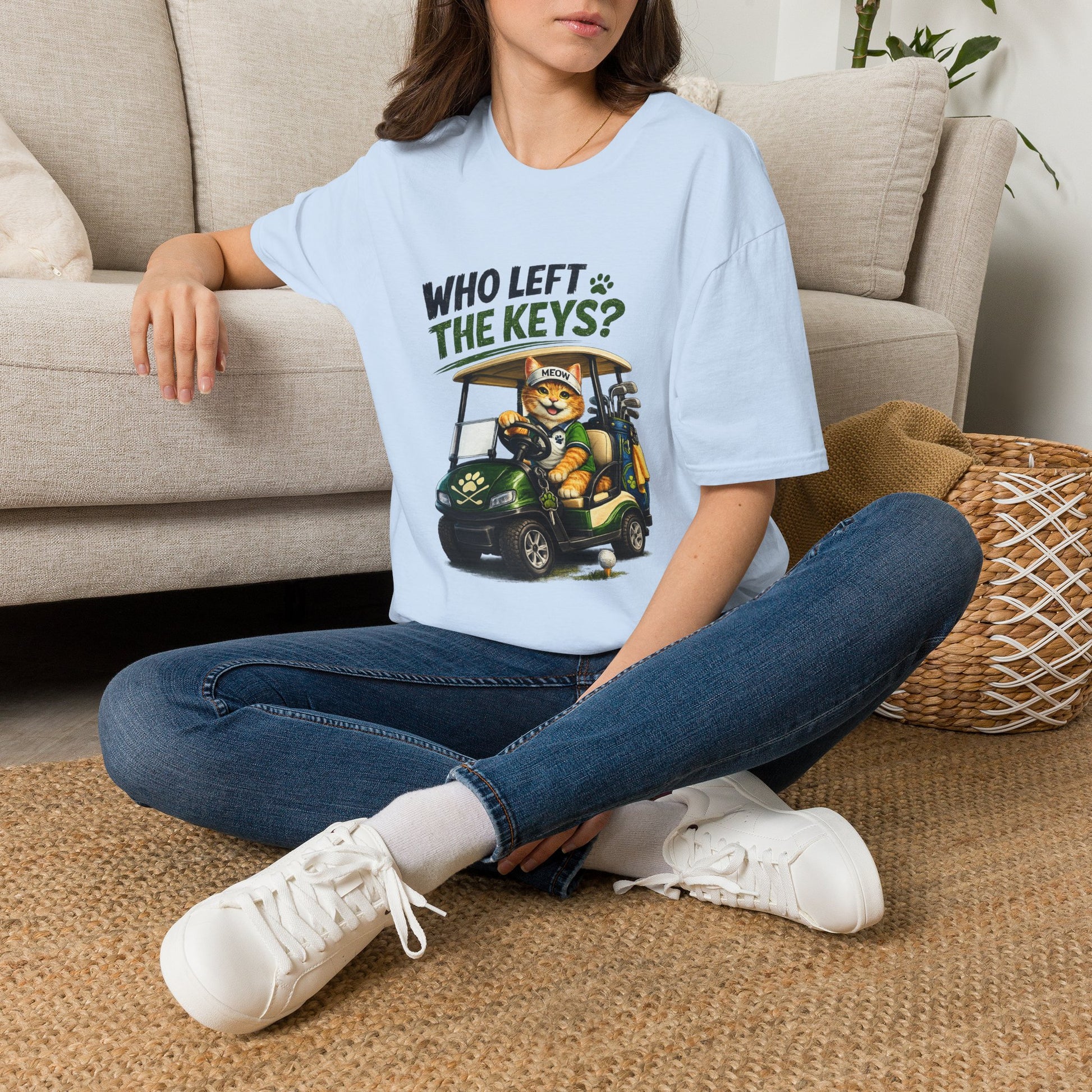 A woman sitting cross-legged on a woven rug in a living room, wearing a light blue graphic t-shirt with a cartoon cat in a golf cart and the text 'WHO LEFT THE KEYS?'.