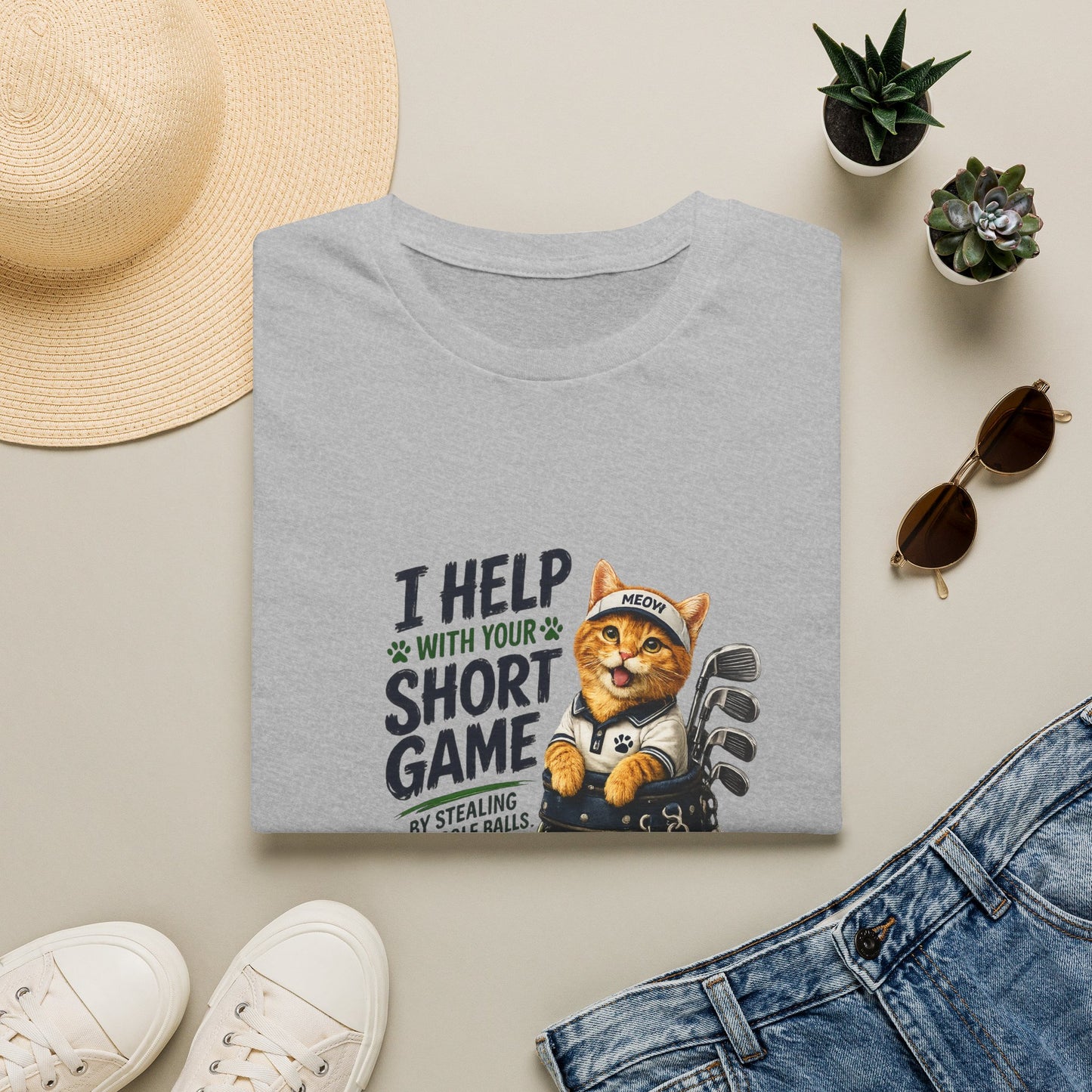 A folded sport grey graphic T-shirt is displayed in a styled flat lay on a light beige surface. The shirt features a cartoon cat wearing a baseball cap and holding golf clubs, with the text 'I HELP WITH YOUR SHORT GAME BY STEALING GOLF BALLS.
