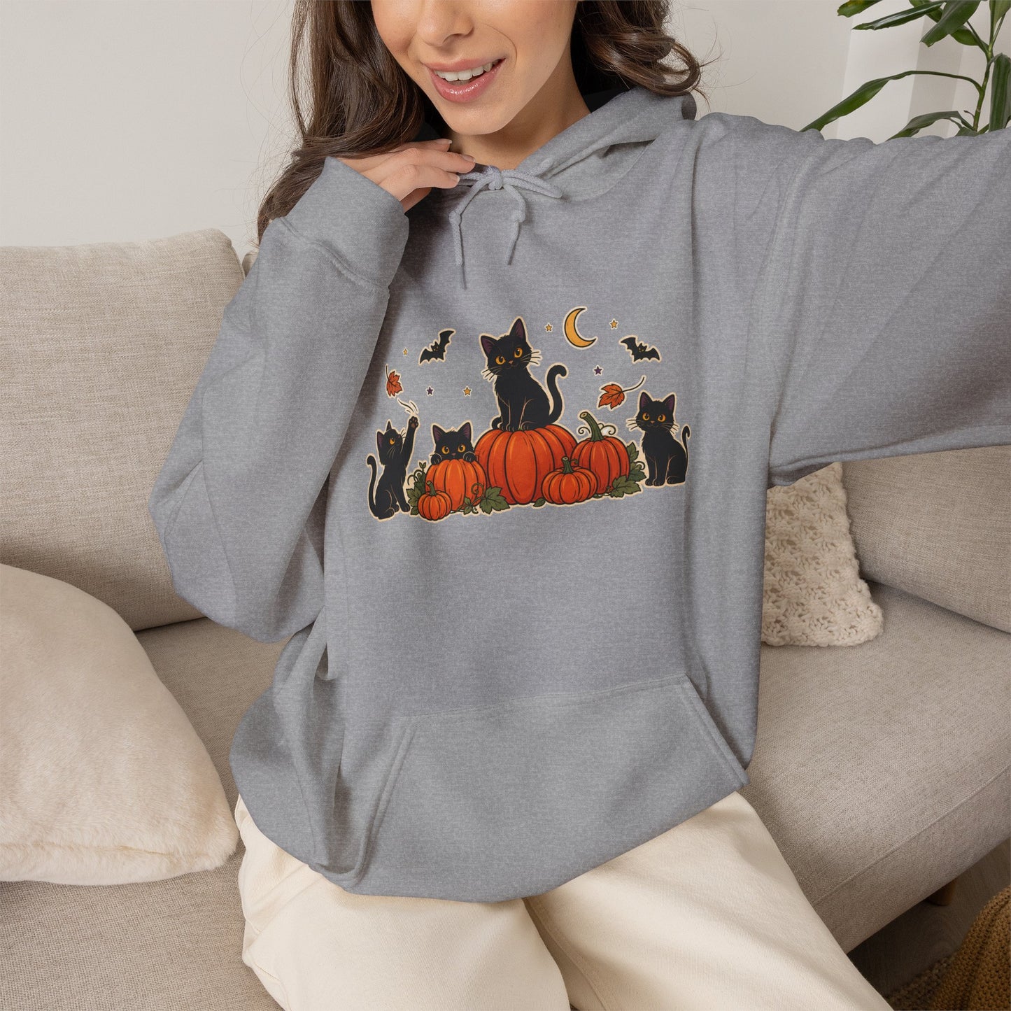A woman wearing a heather gray pullover hoodie with a Halloween graphic of black cats sitting on orange pumpkins, with bats, a crescent moon, and falling leaves. She is sitting on a beige couch.