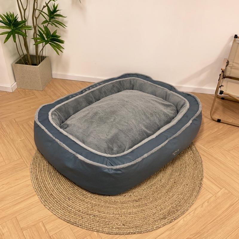Gray pet bed on a wooden floor with a plant and chair in the background