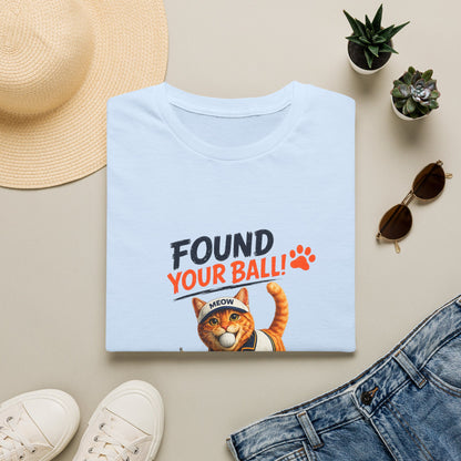 A folded light blue T-shirt with a graphic print reading 'FOUND YOUR BALL!' in black and orange text, plus an orange cat illustration, shown in a styled flat lay with a straw hat, small potted succulents, sunglasses, white sneakers, and blue jeans on a beige surface.